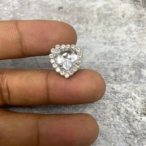 May include: A clear, heart-shaped rhinestone surrounded by a halo of smaller, clear rhinestones. The heart-shaped gem is set in a silver-colored metal. The jewelry piece is a close-up shot.
