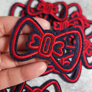 May include: Navy blue and red embroidered bow patches. The patches feature a bow design with a red outline and a navy blue interior. The patches are made of a felt-like material and are suitable for various craft projects.