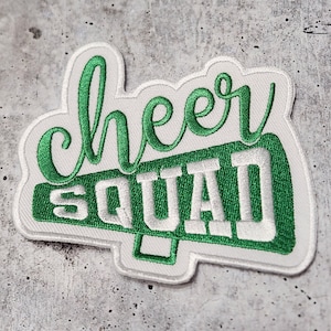Puede incluir: Parche blanco para planchar con texto bordado en verde que dice "cheer squad" dentro de una forma de megáfono verde.