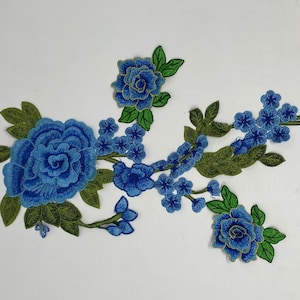 May include: Embroidered floral applique featuring a large blue rose and smaller blue flowers with green leaves. The applique has gold trim and is set against a white background. This decorative patch is ideal for clothing or craft projects.