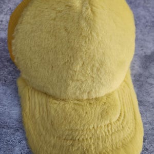 May include: A yellow fuzzy trucker hat with a mesh back. The hat has a curved brim and is made of a soft, plush material.