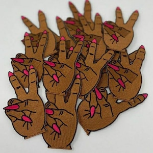 May include: A set of brown fabric patches featuring a hand making the peace sign with pink nail polish.