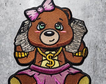 Puede incluir: Un oso de peluche marrón con un lazo rosa, una cadena de oro con un dije de signo de dólar y sosteniendo dos fajos de dinero. El oso lleva una falda rosa y tiene ojos verdes.