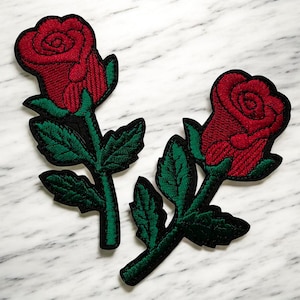 Peut inclure: Deux écussons brodés en forme de rose. Chaque écusson présente une rose rouge vibrante avec des coutures détaillées, des feuilles vertes et une bordure noire. Les écussons sont conçus pour être cousus sur des vêtements ou des accessoires.