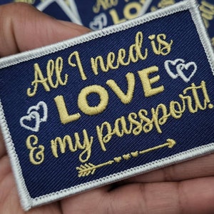 May include: A navy blue embroidered patch with white stitching around the edges. The patch features gold embroidery that reads "All I need is LOVE & my passport!" with two white hearts and an arrow pointing to the right.