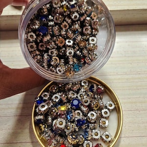 Peut inclure: Un pot en plastique transparent et un plat à bord doré remplis de petites perles en forme de fleur en métal argenté. Les perles ont des centres de pierres précieuses colorées et à facettes dans les tons de bleu, rouge, jaune et transparent. Les perles sont éparpillées sur une surface claire veinée de bois.