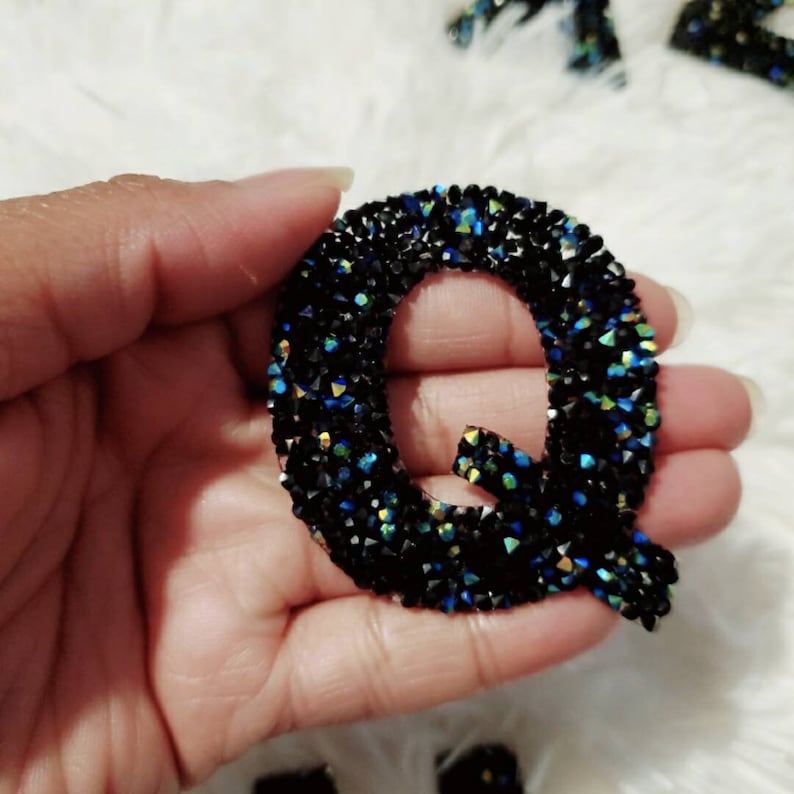Peut inclure: Une lettre Q noire en strass iris&eacute;s. La lettre est tenue dans une main.