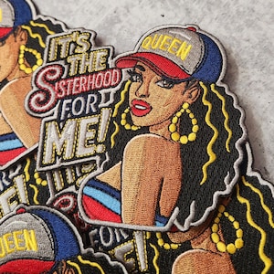 May include: A colorful embroidered patch featuring a woman with long black hair wearing a baseball cap with the word "QUEEN" on it. The patch has the text "It's the Sisterhood for Me!" in a stylized font.