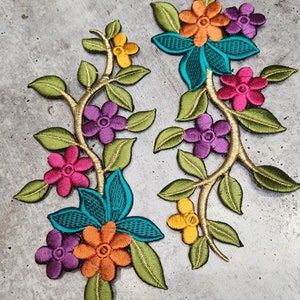 Peut inclure: Deux appliques florales brodées. Chaque applique présente une tige dorée avec des feuilles vertes et des fleurs colorées dans les tons orange, rose, violet, jaune et turquoise. Les appliques sont probablement destinées à des vêtements ou à des projets d'artisanat.