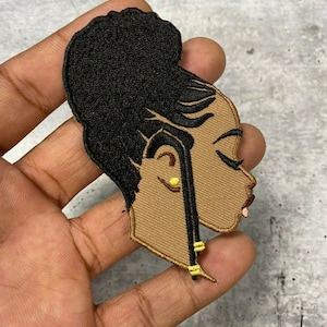 May include: Embroidered iron-on patch featuring a brown-skinned woman with a black bun hairstyle and gold earrings. The woman is looking to the side and has a neutral expression.
