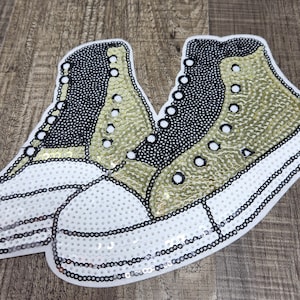 May include: A pair of white, black, and gold sequined sneakers. The sneakers are embroidered with black stitching and have a white sole.