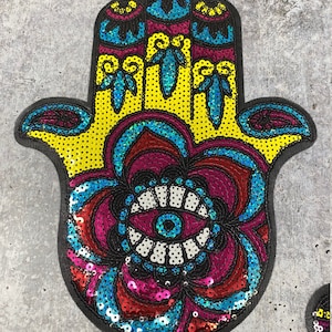 以下が含まれることがあります： 中央に大きな目があるカラフルな手の形のワッペン。ワッペンはスパンコールでできており、黒い背景があります。手は黄色、青、ピンクで、目は青、白、黒です。