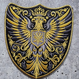 Peut inclure: Un écusson brodé noir et or représentant un aigle héraldique aux ailes déployées, une couronne et un écu orné d'un motif floral stylisé.