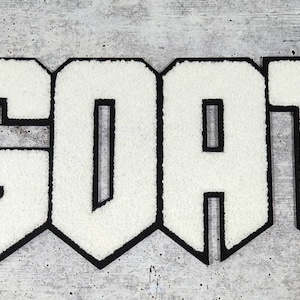 Può includere: Toppa in feltro bianco con la scritta "GOAT" in grassetto, con contorno nero. La toppa ha un aspetto strutturato e sfocato ed è posta su uno sfondo grigio screziato.
