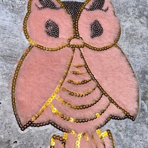 May include: A pink owl applique with gold sequins. The owl has a fluffy texture and is decorated with gold sequins.