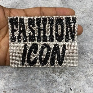 May include: A rectangular, bedazzled accessory with the words "FASHION ICON" spelled out in black and silver rhinestones. The background is a light gray, textured surface.