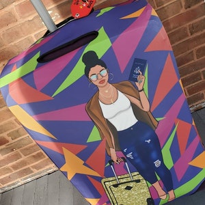 Puede incluir: Una funda de equipaje de color morado, verde, naranja y amarillo con una ilustraci&oacute;n de dibujos animados de una mujer que lleva una chaqueta marr&oacute;n, una camisa blanca, unos vaqueros azules y gafas de sol. La mujer lleva un pasaporte y una maleta dorada. La funda de equipaje tiene una etiqueta que dice "World Traveler" y "Black Owned".