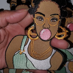 May include: Embroidered patch featuring a cartoon illustration of a Black woman with brown skin, wearing glasses, a green tank top, and gold hoop earrings. She is blowing a pink bubble with her mouth.