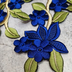 May include: Blue embroidered floral applique with green leaves. The flowers are in a variety of sizes and shapes, and the leaves are outlined in gold thread.