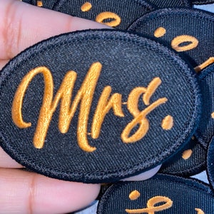 May include: A black fabric patch with a gold embroidered word "Mrs." in a cursive font.