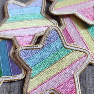 May include: Embroidered star patches in pastel rainbow colors. Each star has a gold border and a white outline. The patches are arranged in a pile, with some overlapping. The patches are a mix of pink, yellow, green, and blue.