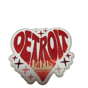 May include: A heart-shaped embroidered patch featuring the word DETROIT in bold red lettering. The design includes a gold skyline silhouette at the base of the heart, surrounded by small red and gold star accents on a white background with a white border.