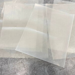 May include: Four sheets of white parchment paper, stacked on top of each other, on a grey textured surface.