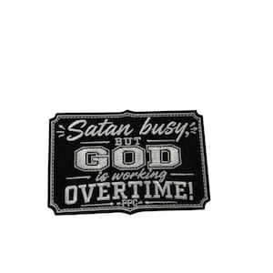 May include: A rectangular black embroidered patch with a white stitched border. The text reads "Satan busy, BUT GOD is working OVERTIME!" in a mix of script and block lettering, with the letters "PPG" centered at the bottom in white thread.