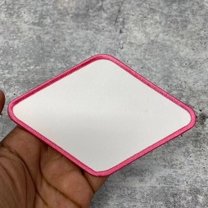 以下が含まれることがあります： ピンクの縁取りのある白いダイヤモンド型のワッペン。ワッペンは布製で、表面が滑らかです。