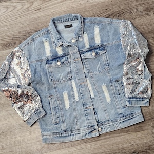 May include: Light blue denim jacket with distressed detailing and silver sequin sleeves. The jacket has a classic collar, button-down front, and multiple pockets. A stylish and unique outerwear piece.