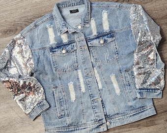 May include: Light blue denim jacket with distressed detailing and silver sequin sleeves. The jacket has a classic collar, button-down front, and multiple pockets. A stylish and unique outerwear piece.