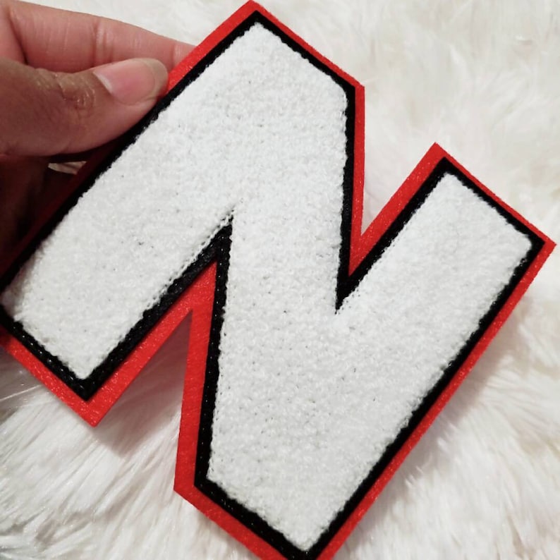 Puede incluir: Letra N de fieltro blanco con contorno rojo y negro. La letra es gruesa y tiene una textura ligeramente en relieve.