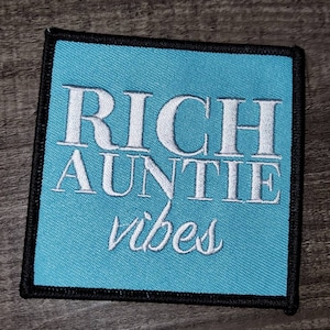 May include: A blue square patch with black stitching around the edges. The patch has white embroidered text that reads "RICH AUNTIE vibes".
