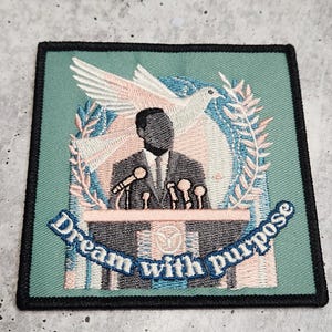 May include: An embroidered patch with a green background featuring a silhouette of a person speaking at a podium with a dove flying above. The text "Dream with purpose" is embroidered in a curved arc below the podium.