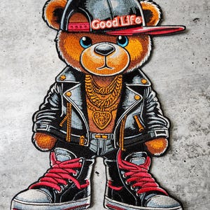 May include: A brown teddy bear wearing a black leather jacket, blue jeans, red sneakers, a gold chain, and a black and red baseball cap with the words "Good Life" on it.
