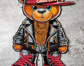 Puede incluir: Un oso de peluche marrón que lleva una chaqueta de cuero negra, pantalones vaqueros azules, zapatillas rojas, una cadena de oro y una gorra de béisbol negra y roja con las palabras "Good Life" en ella.
