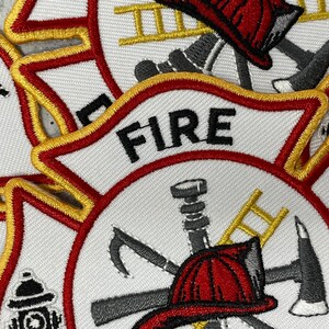 Op de afbeelding: Close-up van een wit geborduurd patch met rode en gouden rand. De patch toont een brandweerhelm, een bijl, een ladder en een brandweerslang. Het woord "FIRE" is in zwarte letters bovenaan geborduurd.