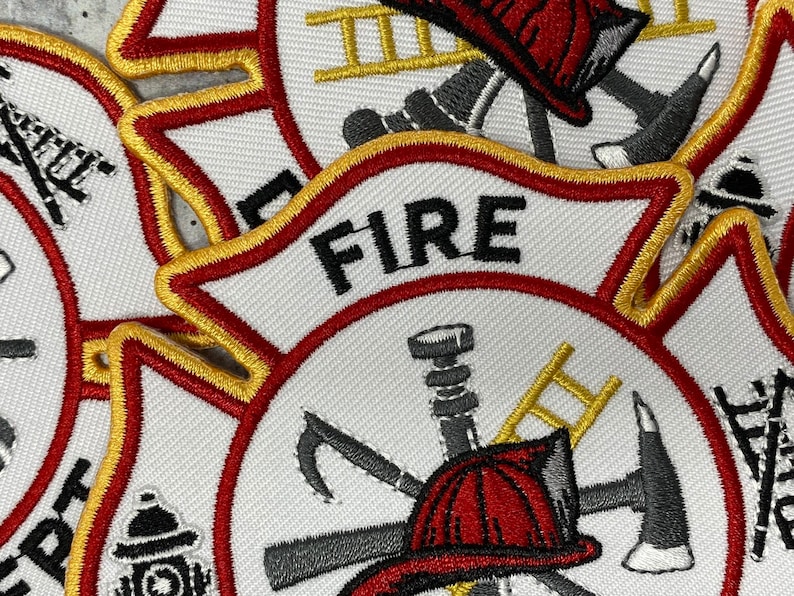 Op de afbeelding: Close-up van een wit geborduurd patch met rode en gouden rand. De patch toont een brandweerhelm, een bijl, een ladder en een brandweerslang. Het woord "FIRE" is in zwarte letters bovenaan geborduurd.