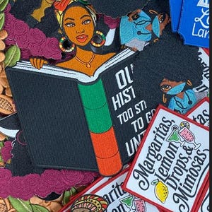 Op de afbeelding: Een verzameling geborduurde patches met diverse ontwerpen. Eén patch toont een zwarte vrouw die een boek leest met de tekst "Our History Too Strong To Go Unt...". Andere patches bevatten cocktail- en inspiratieontwerpen.