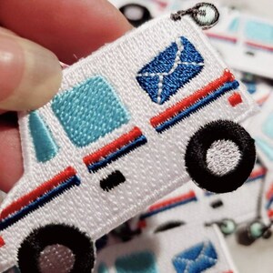 Pu&ograve; includere: Toppa da stirare ricamata bianca di un camion postale con una busta blu, strisce rosse e blu e ruote nere.
