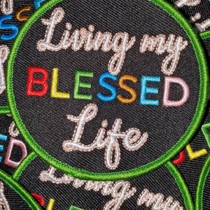 May include: Close-up of a black fabric patch with green stitching around the edges. The patch says "Living my BLESSED Life" in a white cursive font. The word "BLESSED" is in rainbow colours.