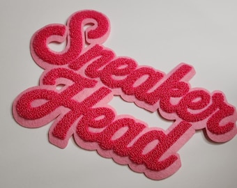May include: A pink and red embroidered patch with the words "Sneaker Head" in a cursive font. The patch has a light pink background and a textured, raised red font. The patch is likely for clothing or accessories.