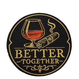 Puede incluir: Un parche bordado circular con fondo negro y borde dorado. Presenta una copa de coñac llena de líquido ámbar, un cigarro encendido con humo y el texto bordado en oro "BETTER TOGETHER" con adornos decorativos en la parte inferior.