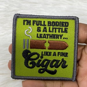 May include: Square patch with a green background and a gray border. The patch features a brown cigar graphic with the text "I'M FULL BODIED & A LITTLE LEATHERY... LIKE A FINE Cigar" in black.