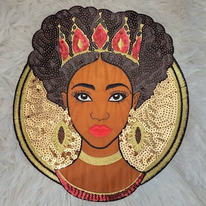 May include: A round, embroidered patch featuring a woman with brown hair, wearing a gold crown and earrings. The background is gold sequins.