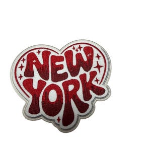 May include: A heart-shaped embroidered patch featuring the text NEW YORK in a bold, red, dripping-style font. The design is outlined in white and grey thread and includes small, scattered four-pointed star accents against a white background.