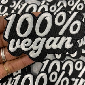 Op de afbeelding: Een close-up van een zwarte stoffen patch met witte geborduurde tekst die "100% vegan" luidt.