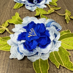 May include: Two blue and white embroidered flower appliques with green leaves and gold accents. The flowers are in a peony style.