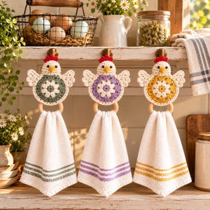 May include: Three hanging kitchen towels, each featuring a crocheted chicken topper with a central flower motif in sage green, lavender, or yellow. The white towels have matching colored stripes at the bottom and are attached to wooden rings.