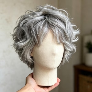 以下が含まれることがあります： シルバーとグレーの髪色が混ざり合った、ショートレイヤーのウィッグ。ふんわりとした質感のウェーブと顔周りを覆う柔らかな前髪が特徴で、明るい色と暗い色の毛束が重なり合い、ボリューム感のある立体的なスタイルを作り出しています。